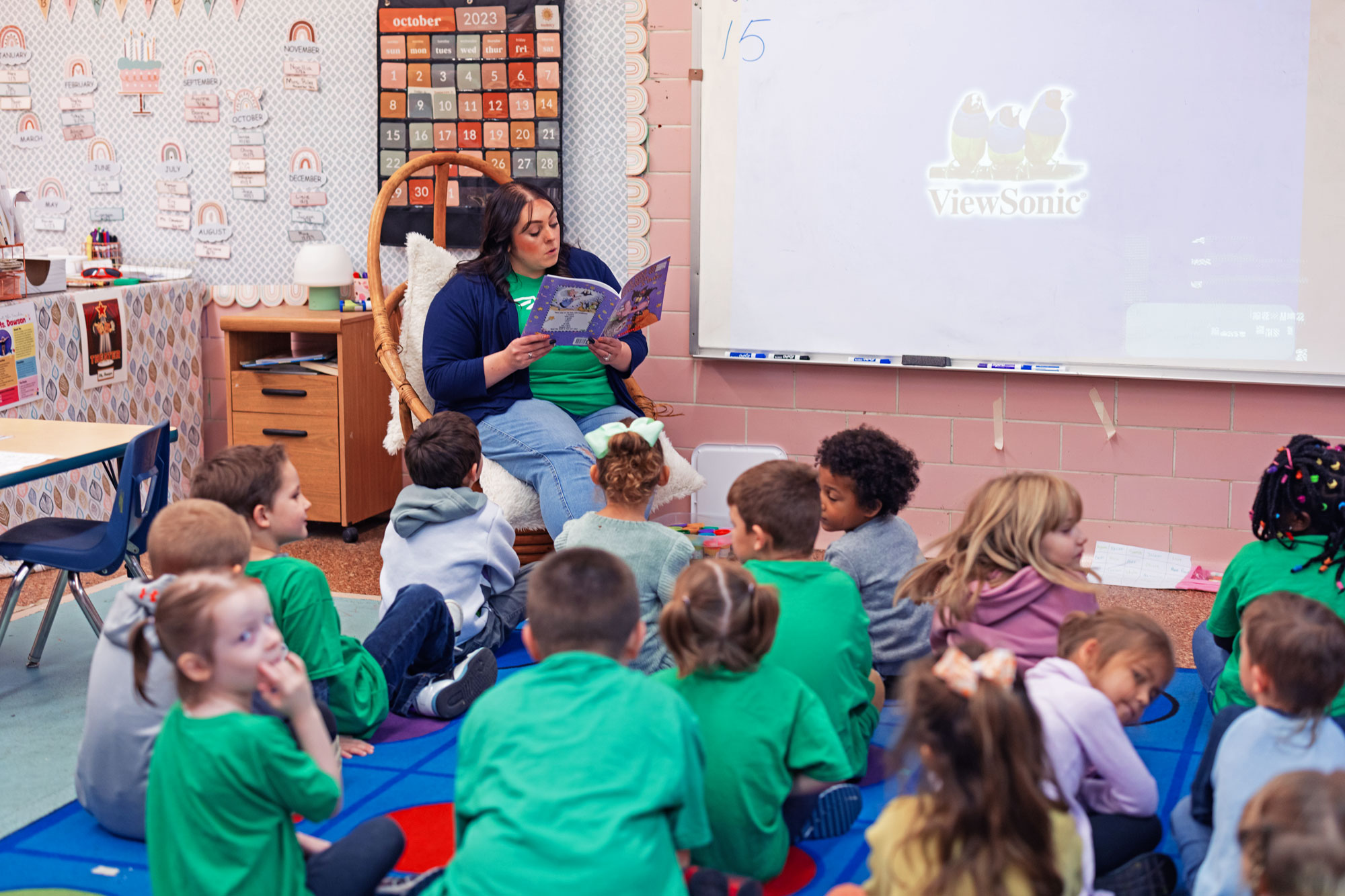 Teacher reads a book aloud to students seated on a classroom rug.