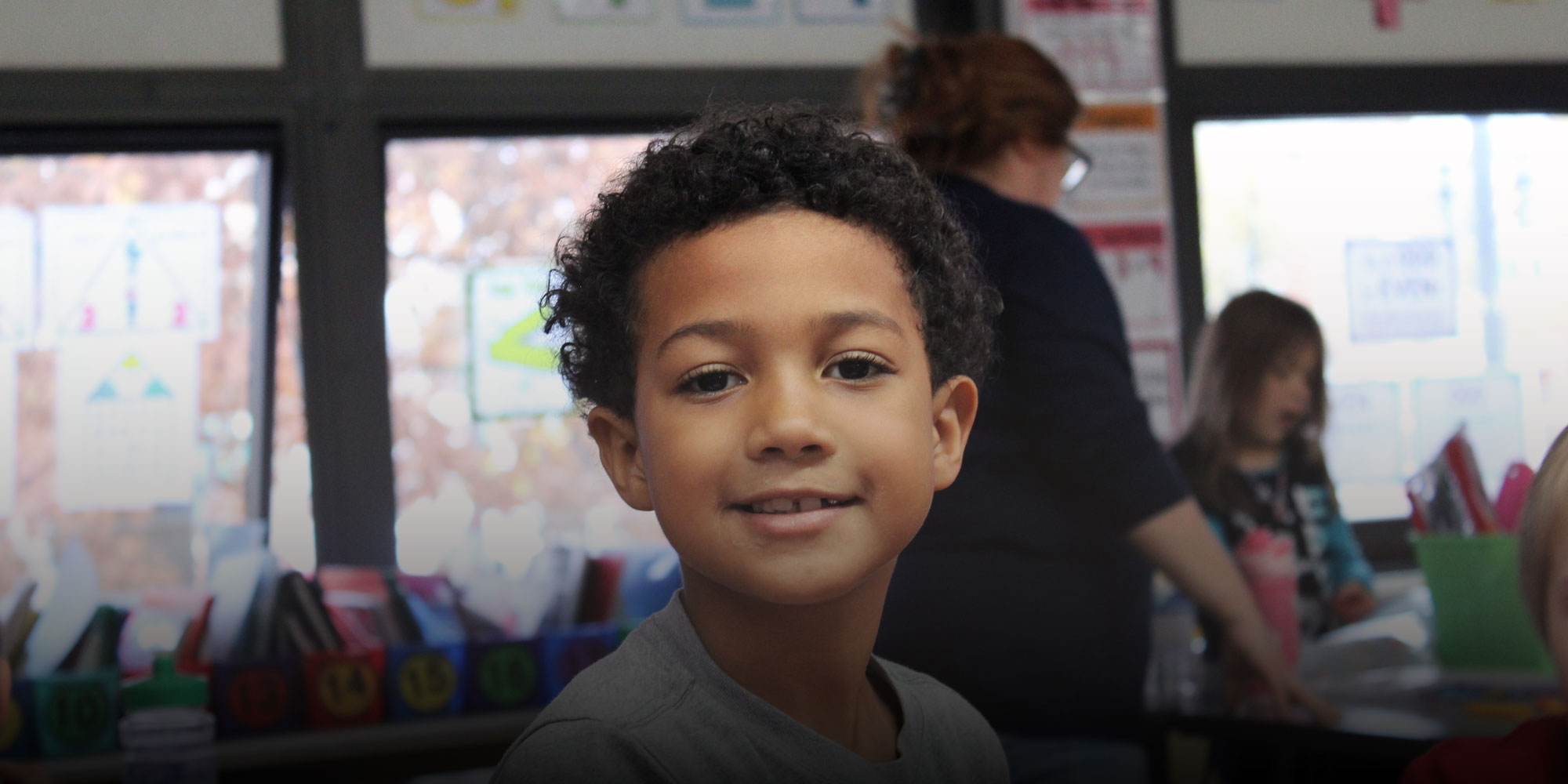 Smiling student in a classroom with a teacher in the background.