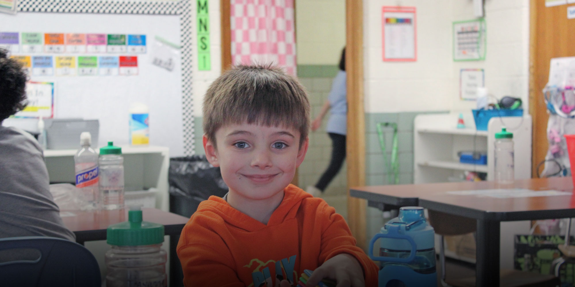 Smiling student in an orange hoodie at a classroom table.