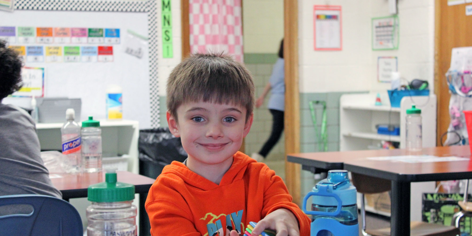 Smiling student in an orange hoodie at a classroom table.